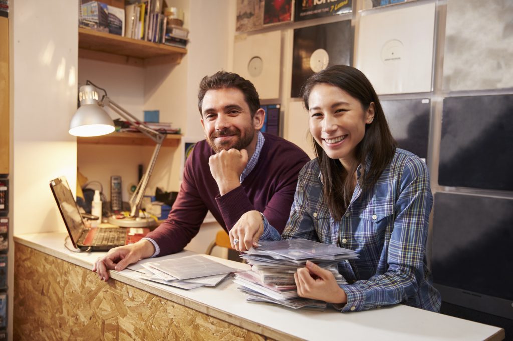 Man and woman working behind the counter at a record shop, smiling to camera